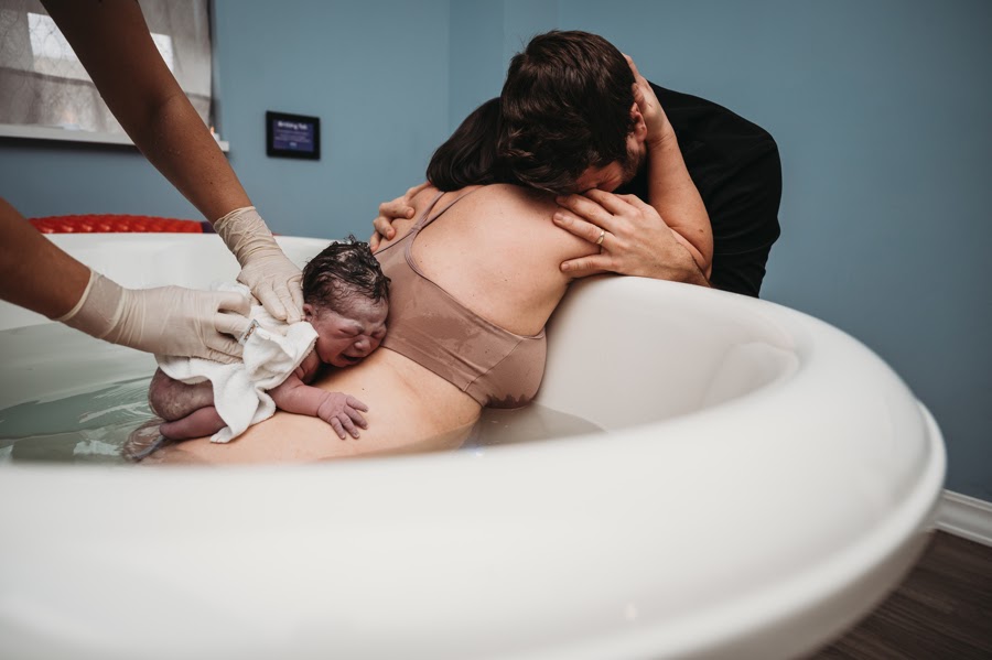 June is placed on Krista's back in the birthing tub. Krista is leaning on the side of the tub and clutching her husband.